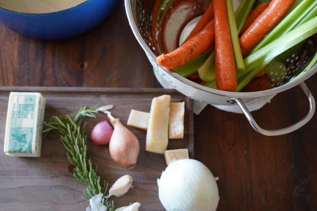 a cutting board with butter, onions and parmesan cheese rinds and next to it is a strainer with vegetables