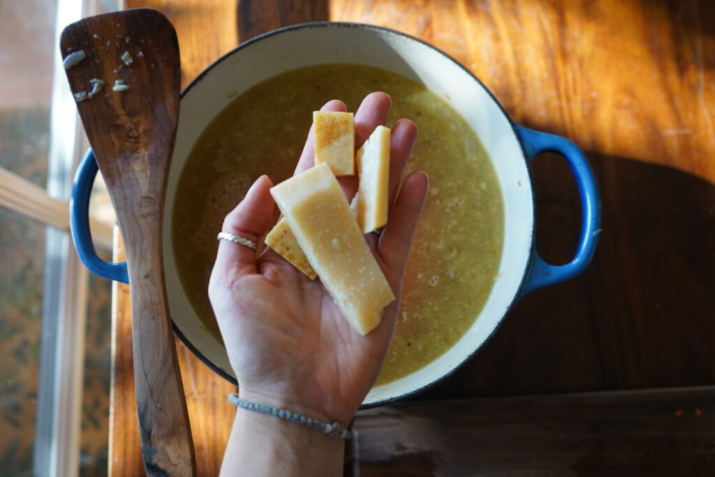 a hand holding parmesan rinds over a dutch oven of soup