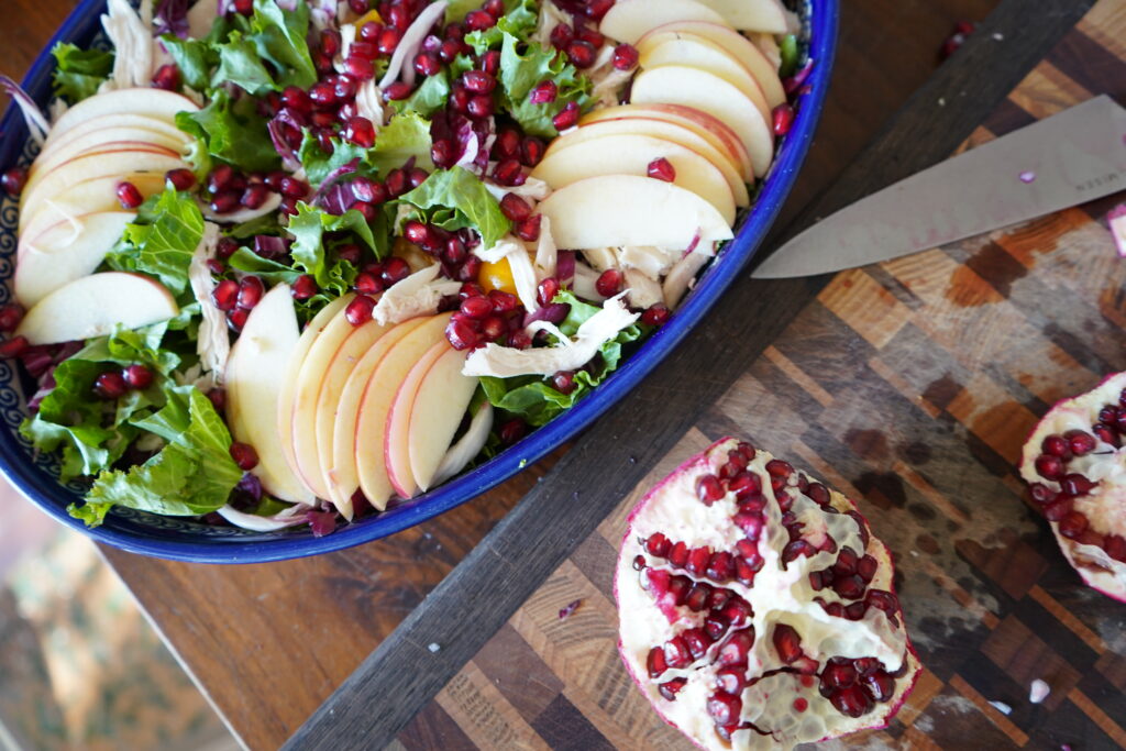 A pomegranate on a cutting board, cut open exposing the seeds and next to it is a salad bowl full of fruits and vegetables.