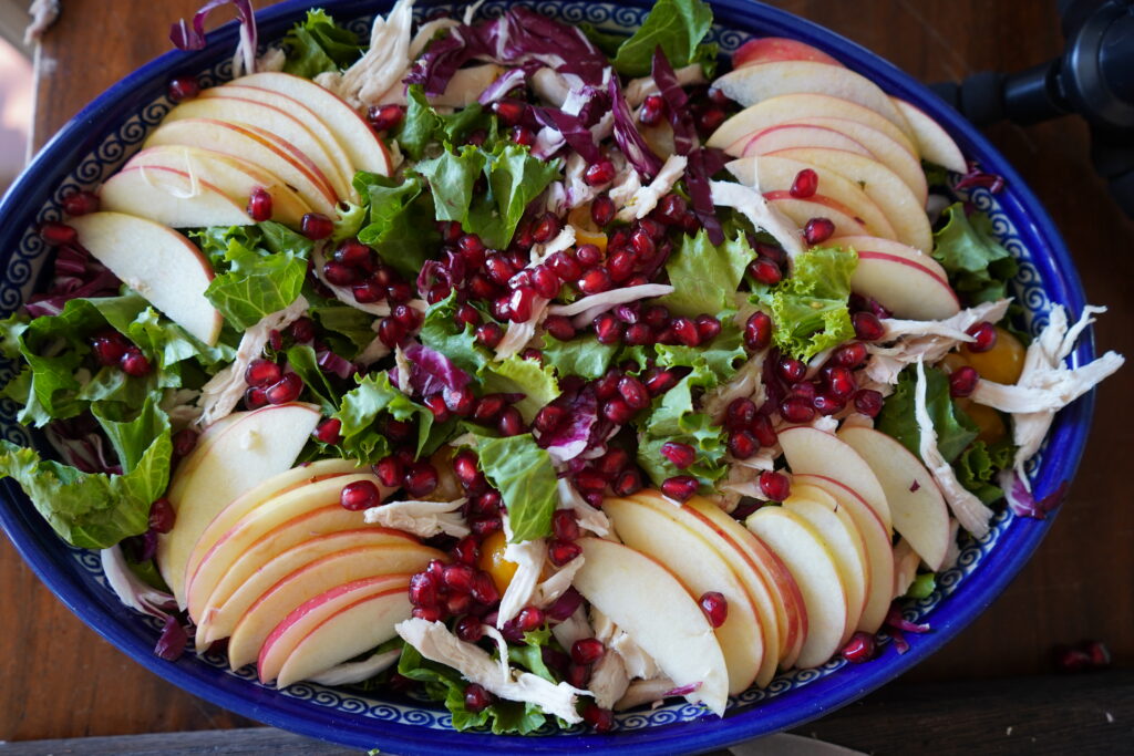 A blue oval salad bowl filled with lettuce, apples, pomegranates and chicken.