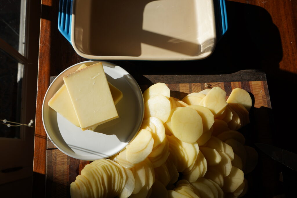 Thinly sliced potatoes on a cutting board, next to a plate of cheese