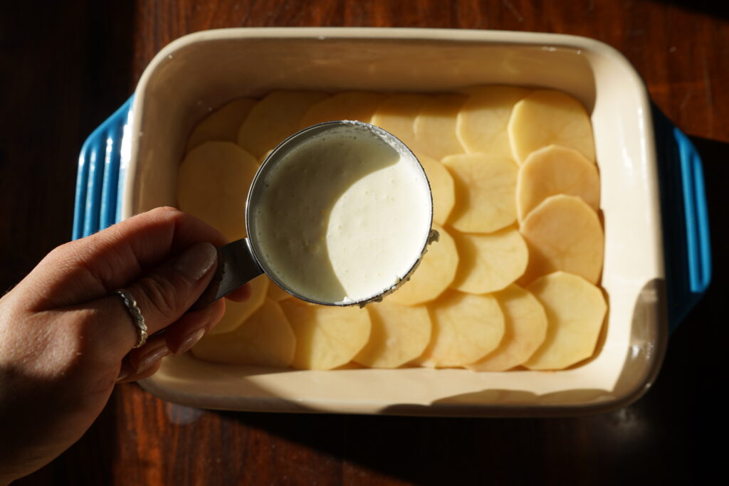 A cup of heavy cream over a baking dish of potatoes