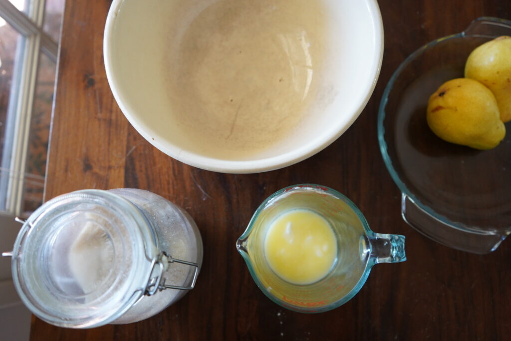 A mixing bowl, 2 pears, a pitcher of butter and a container of sugar