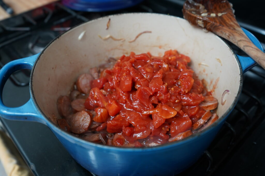 tomatoes being added to a pot of sausages