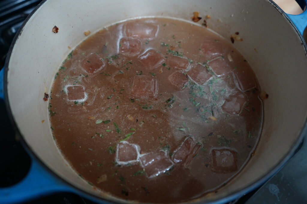 frozen broth being added to a Dutch oven off herbs and roasted garlic