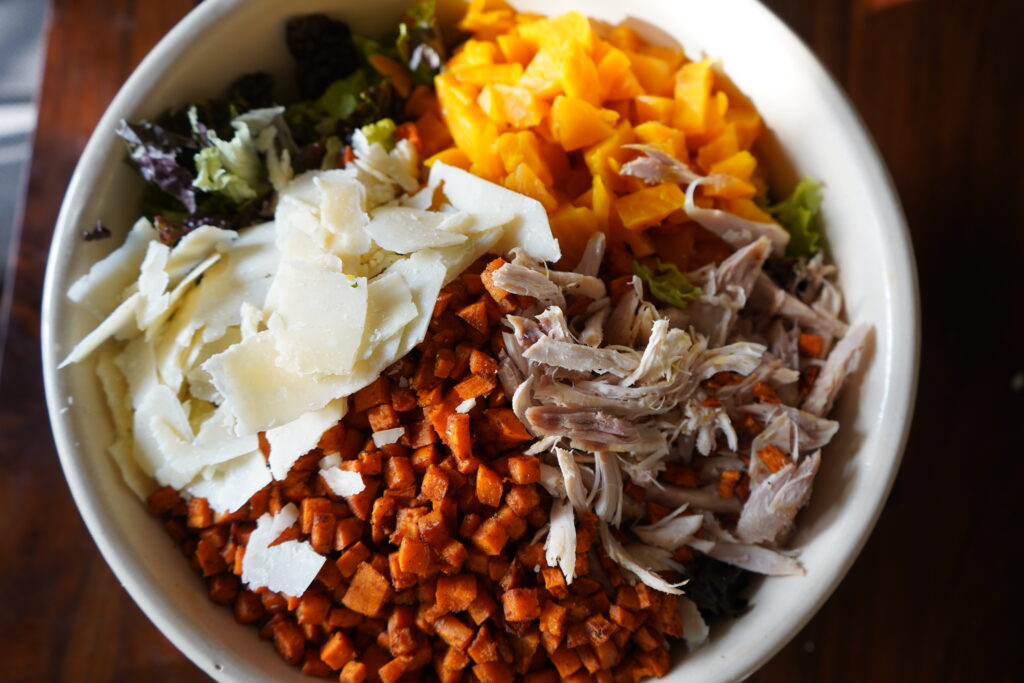 a huge salad in a bowl topped with parmesan, chicken and sweet potato