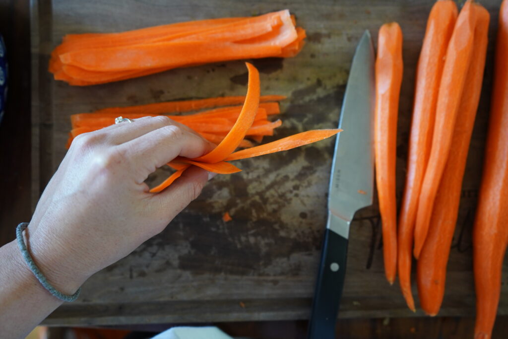 a hand holding thinly sliced carrot and whole carrots sitting on a cutting board