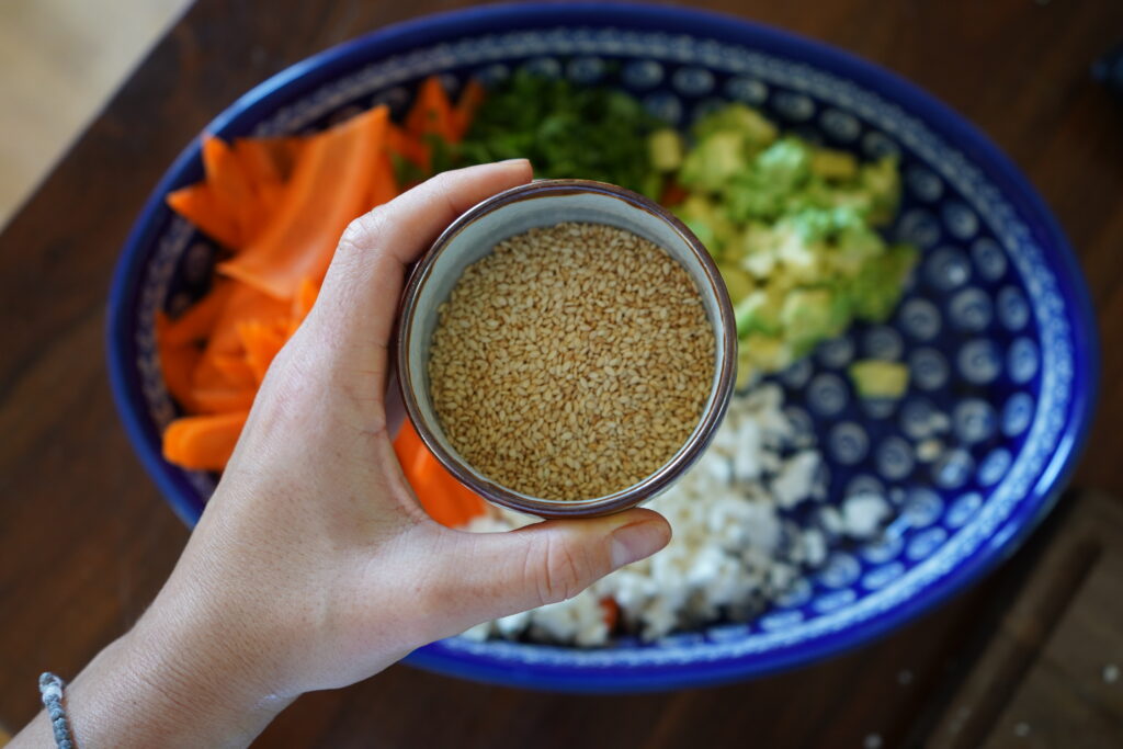 a hand holding a little bowl of toasted sesame seeds over a bowl of salad