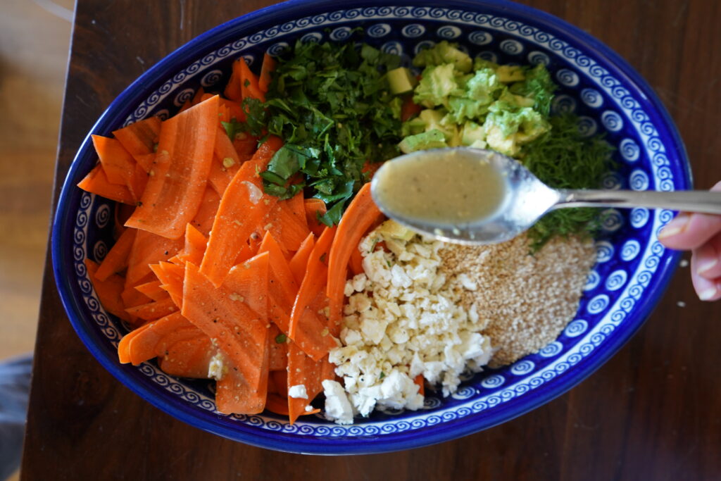 a blue oval salad bowl with a carrot salad inside and a silver spoon pouring dressing on top