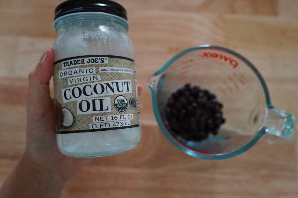 a hand holding coconut oil over a glass dish of chocolate chips