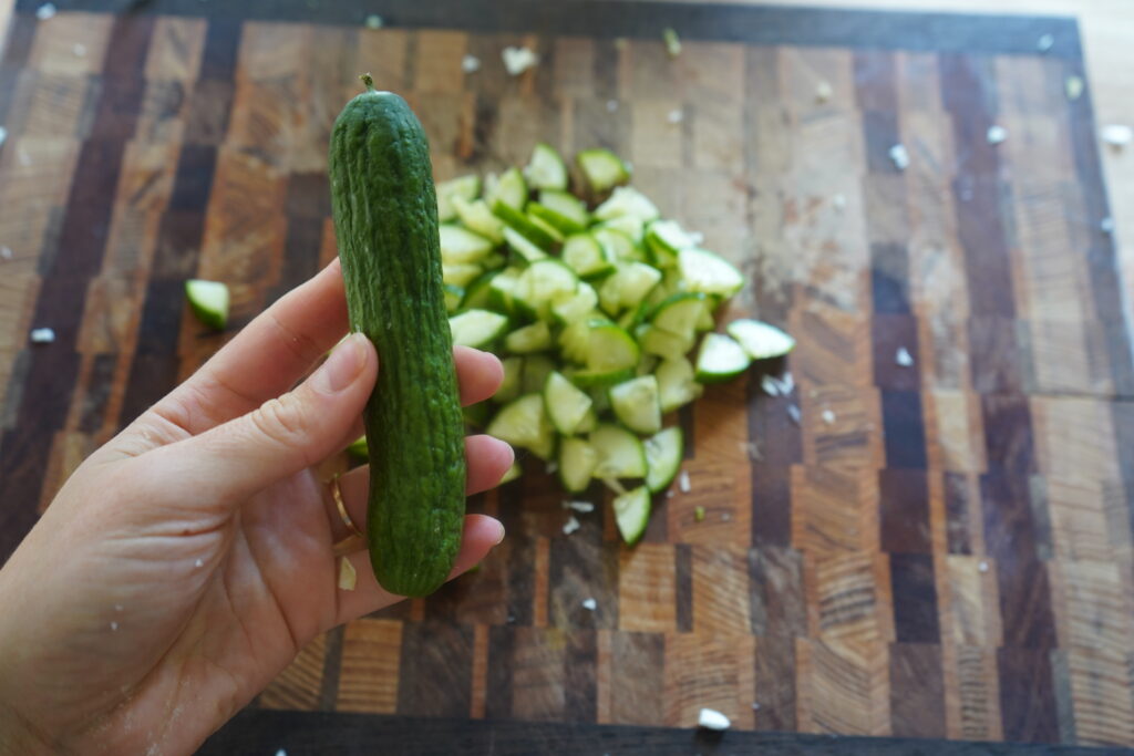 a hand holding a cucumber and chopped cucumber on a cutting board