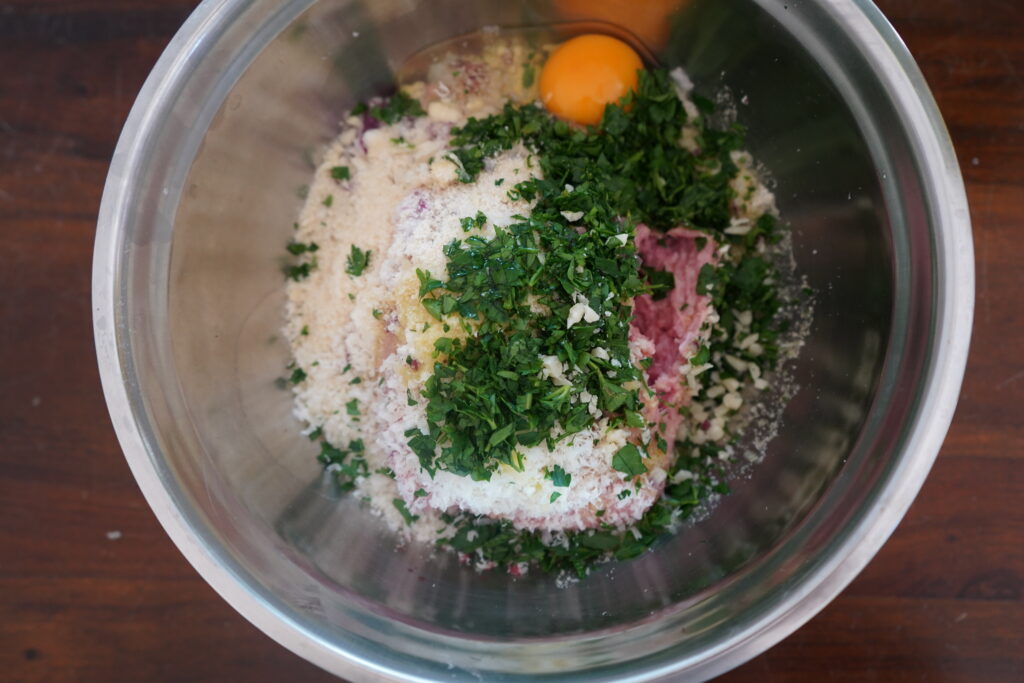 an egg added to a mixing bowl of beef and onions and parsley