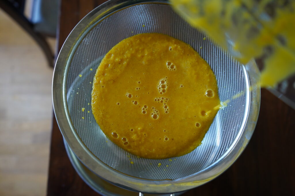 chunky orange and turmeric juice being poured through a strainer