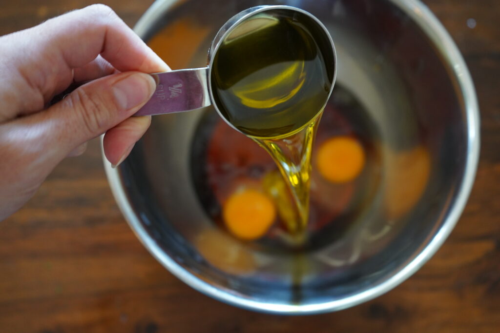 olive oil being poured into a mixing bowl of eggs and maple syrup