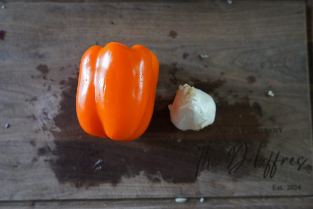 a orange bell pepper and a head of garlic on a brown cutting board
